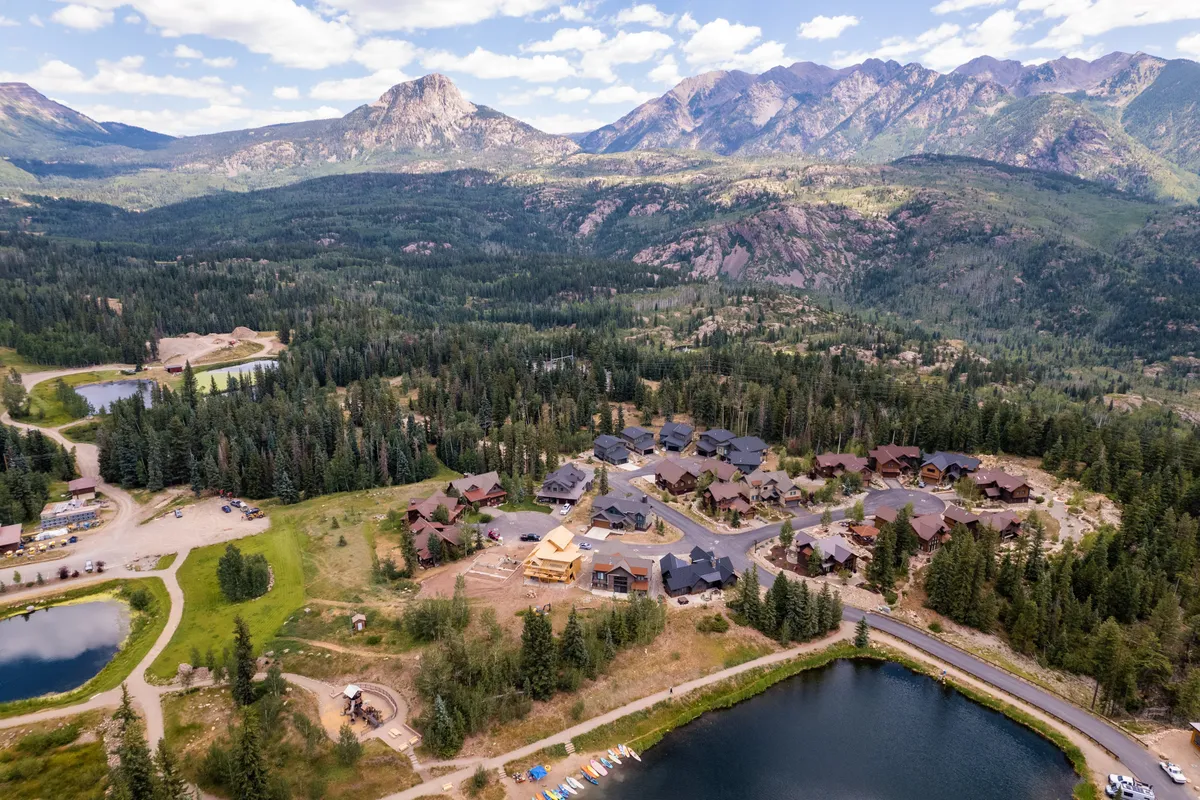 An inviting exterior landscape featuring a meandering pond, walking pathways, a distant tree line, and a neighboring residential area, all framed by dramatic mountain peaks. The open space suggests easy access to nature and outdoor exploration.