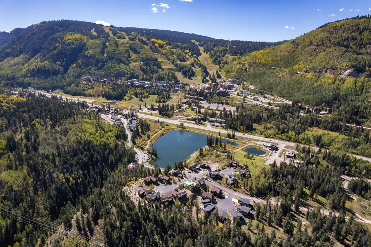 A panoramic exterior view capturing the tranquil lake bordered by lush forested areas, with mountain slopes rising in the background and residential buildings nestled nearby. The serene water, open sky, and rugged mountains evoke the adventure and relaxation promised by Colorado summers.
