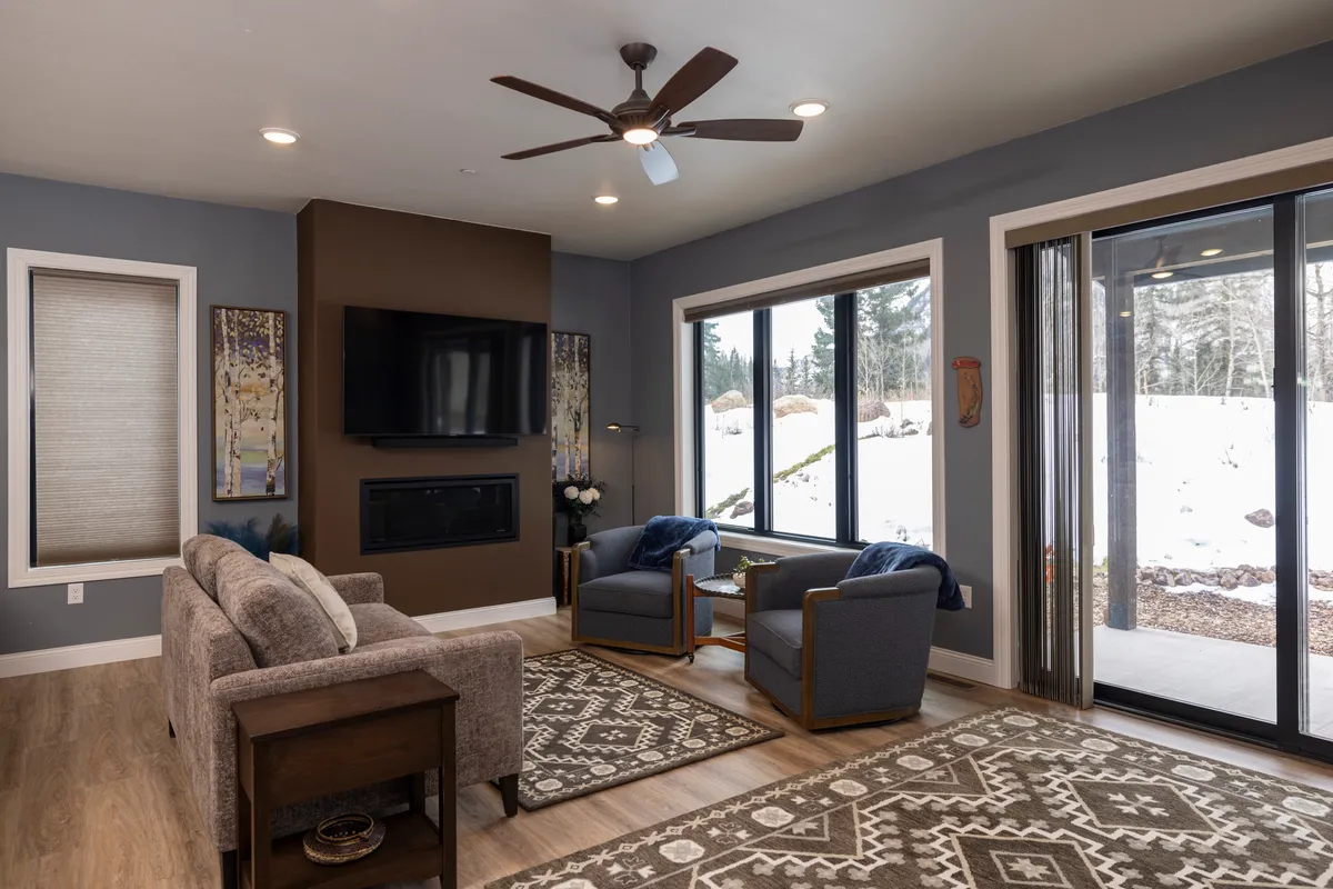 A welcoming living area with a comfortable sofa, wooden armchairs, a crackling fireplace, large windows framing the mountain view, and thoughtfully selected wall art. The space feels cozy yet expansive, ideal for relaxing or socializing.