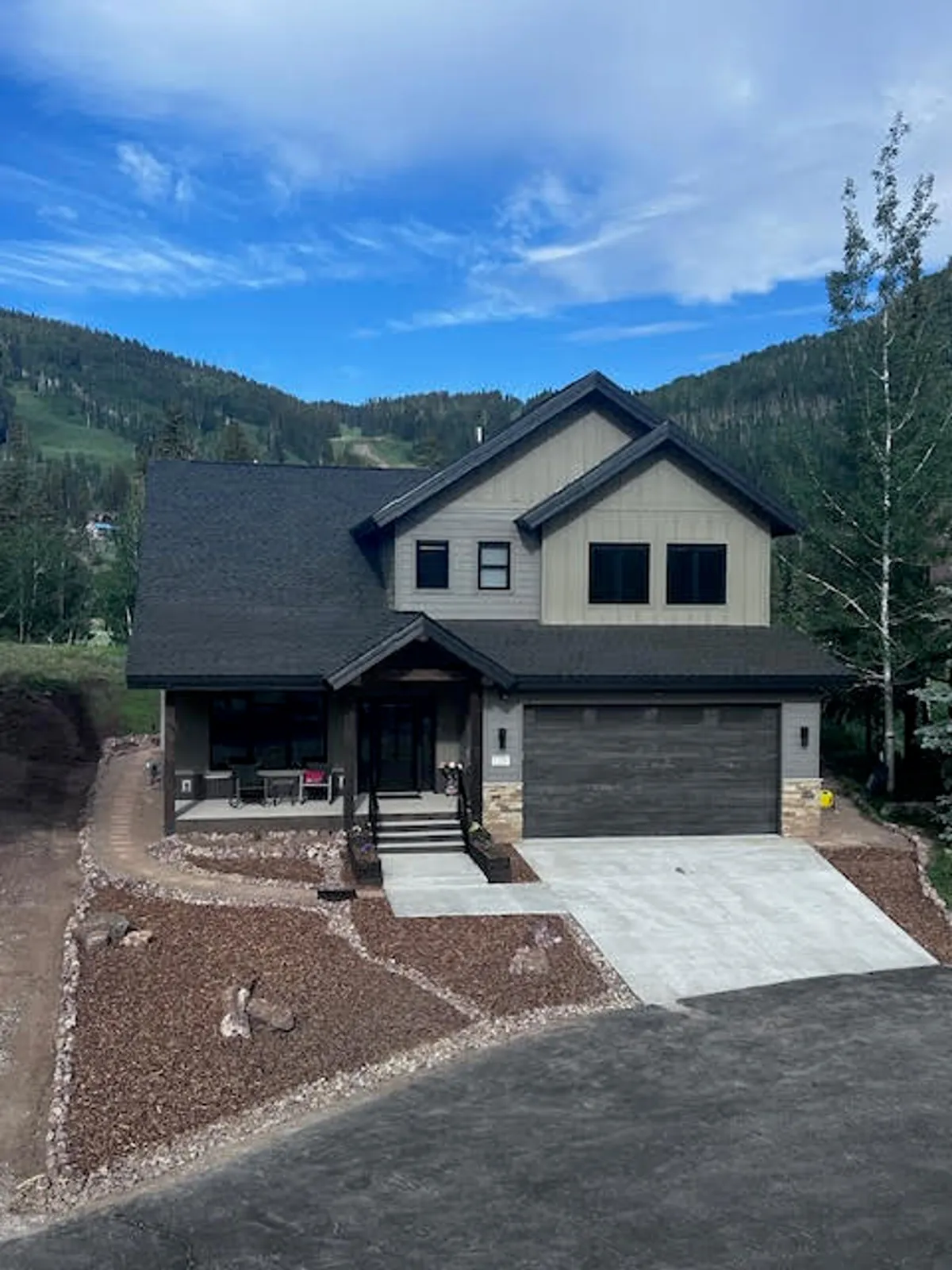 A wide exterior shot of the home showcases its modern architectural design, the welcoming front porch, the landscaped garden, and the driveway, with a backdrop of mountain scenery. The scene exudes serenity and hints at outdoor relaxation and mountain views.