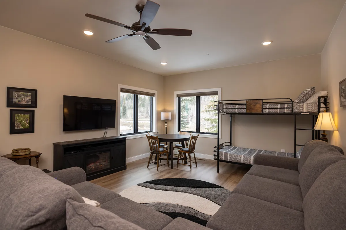 A cozy recreation room featuring a plush sectional sofa facing a television, a dining/game table, and large windows that flood the space with natural light. The room is arranged to support both family gatherings and leisure activities like relaxing, playing games, or watching movies together.