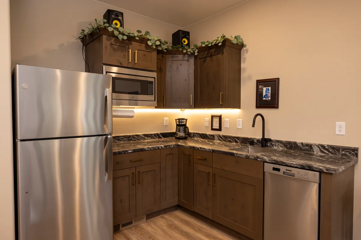 A compact bar kitchenette with stainless steel appliances, dark wood cabinets, marble countertop, and a coffee maker. The area is well-lit and convenient for preparing snacks or drinks during gatherings.