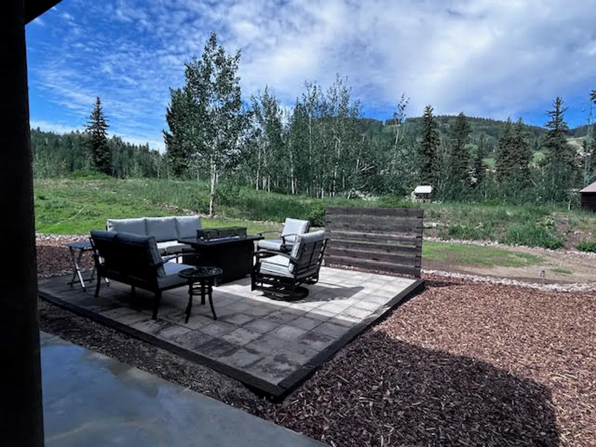 A cozy, elevated outdoor seating area with a wooden privacy screen, surrounded by lush greenery, perfect for relaxing, socializing, or enjoying nature. The scene suggests a tranquil spot for watching sunsets over the mountains or hosting outdoor meals with friends and family.