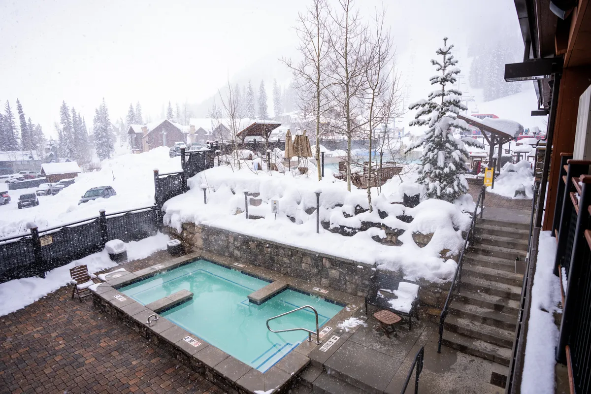 An outdoor hot tub sits nestled in a snow-covered landscape, surrounded by trees and accessed by wooden steps. Steam rises from the bubbling water, inviting guests to soak and enjoy the picturesque winter views.