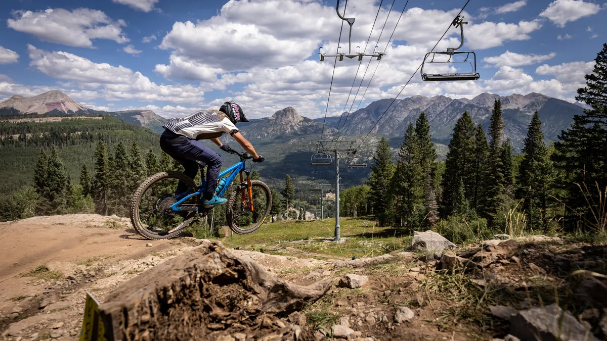 A dynamic exterior shot capturing a mountain biker riding near a ski lift, framed by a majestic mountain range and pine trees. The image suggests year-round adventure and stunning scenery.