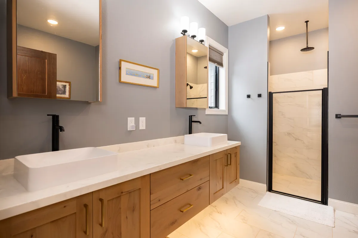 A modern bathroom with double sinks, a large mirror, a shower enclosure, and wooden cabinetry. The space is airy and bright, designed for both functionality and relaxation.