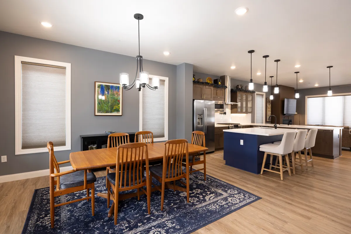 A warm and welcoming interior scene featuring a spacious dining table, elegant chandelier, kitchen island, and visible fridge. The space is arranged for both family meals and entertaining, with artwork adding a homey touch.